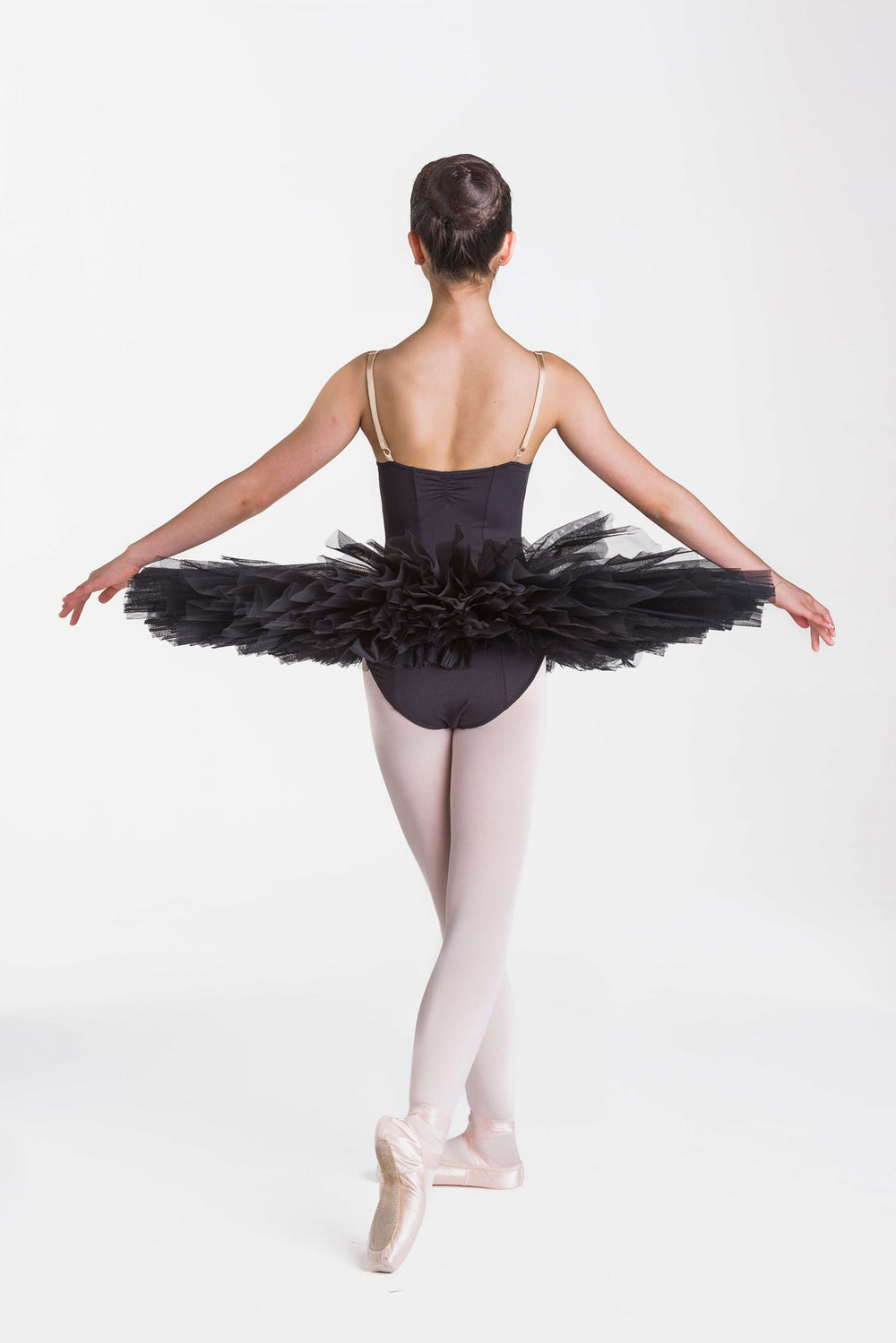 A young ballerina viewed from behind, wearing a black tutu with thin spaghetti straps, black tights, and pink pointe shoes, posed against a seamless white studio background.