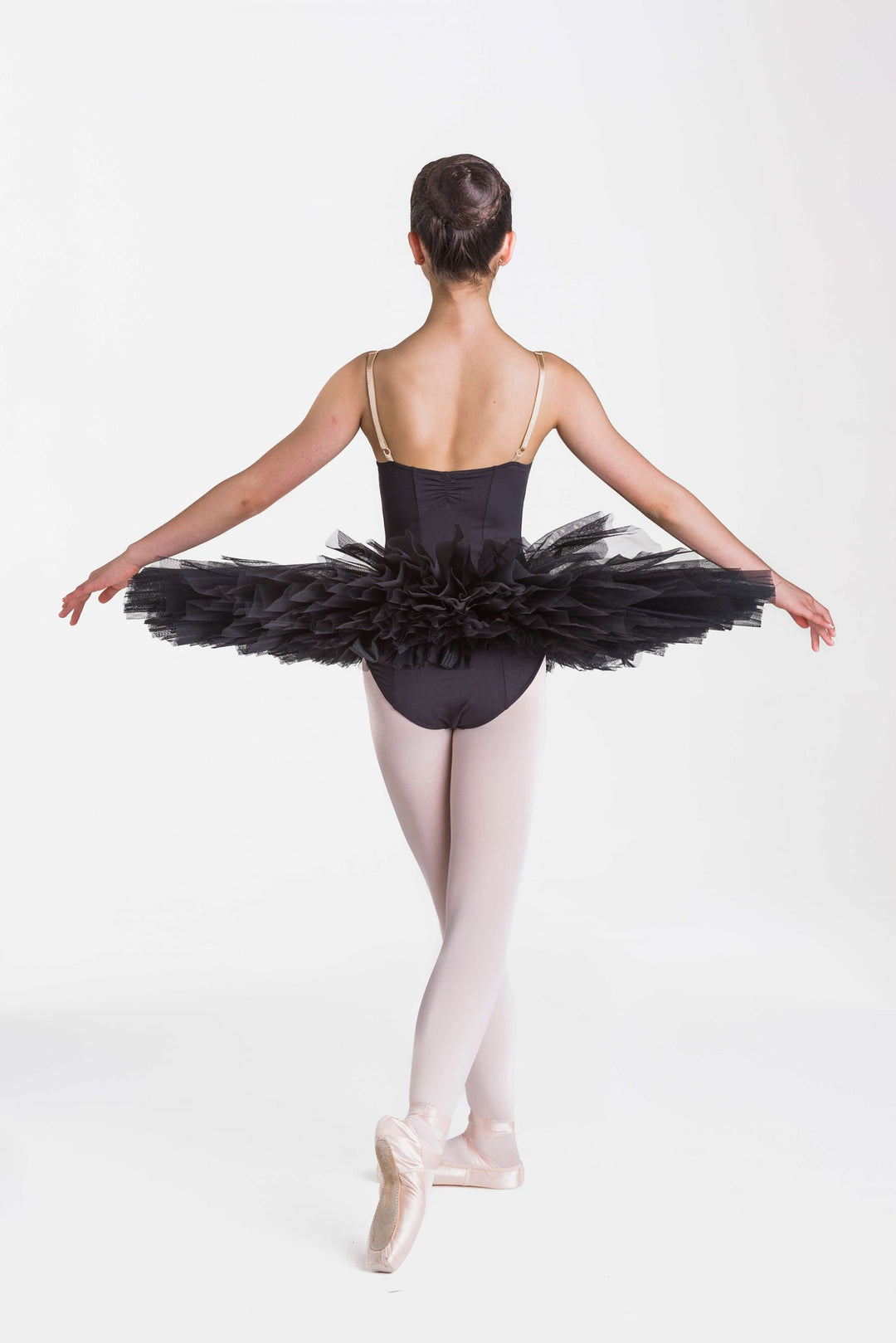A young ballerina viewed from behind, wearing a black tutu with thin spaghetti straps, black tights, and pink pointe shoes, posed against a seamless white studio background.