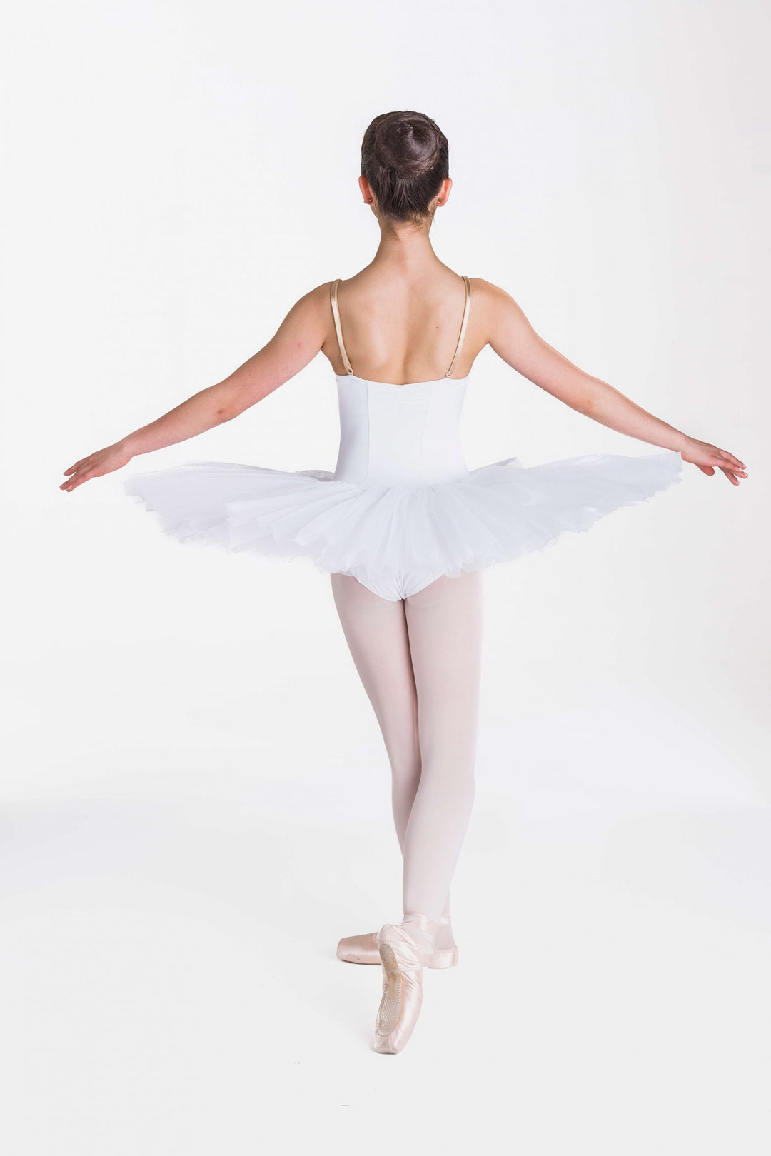 A young ballerina viewed from behind, wearing a white tutu, white bodice, and light pink tights, posing en pointe against a seamless white studio background.