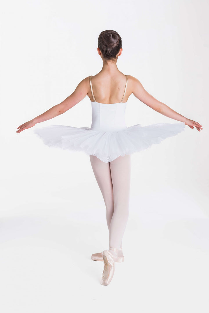 A young ballerina viewed from behind, wearing a white tutu, white bodice, and light pink tights, posing en pointe against a seamless white studio background.