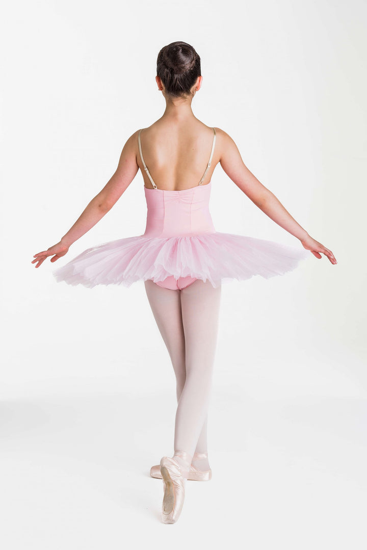 A young ballerina viewed from behind, wearing a light pink tutu, pale pink leotard, white tights, and pointe shoes against a plain white studio background.