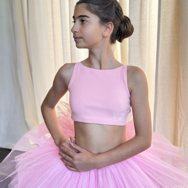 Young girl in a pink ballet outfit standing against a neutral curtain background